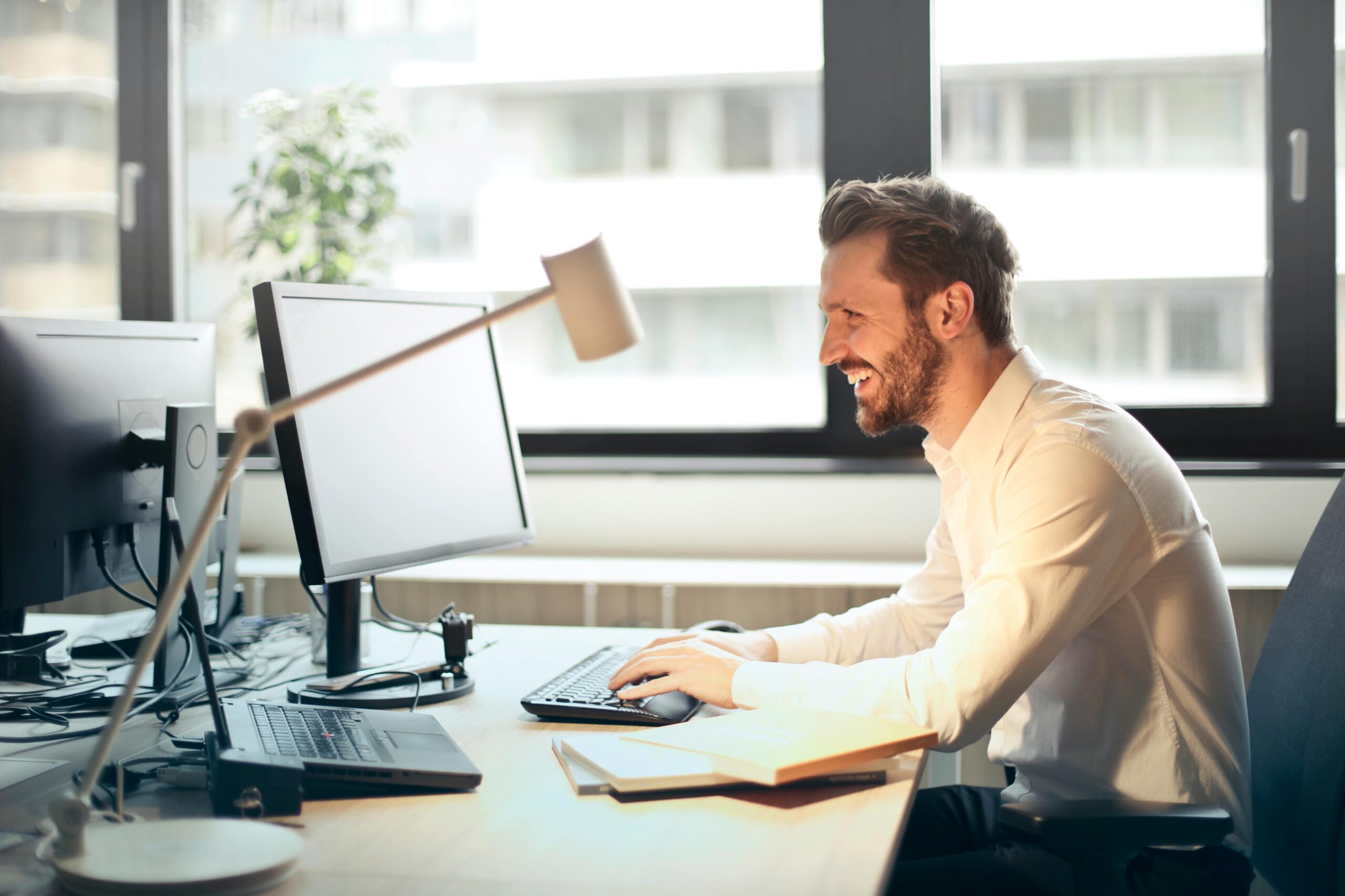 Online Classes A man smiling while working at an office desk with a computer and natural daylight streaming in through large windows.