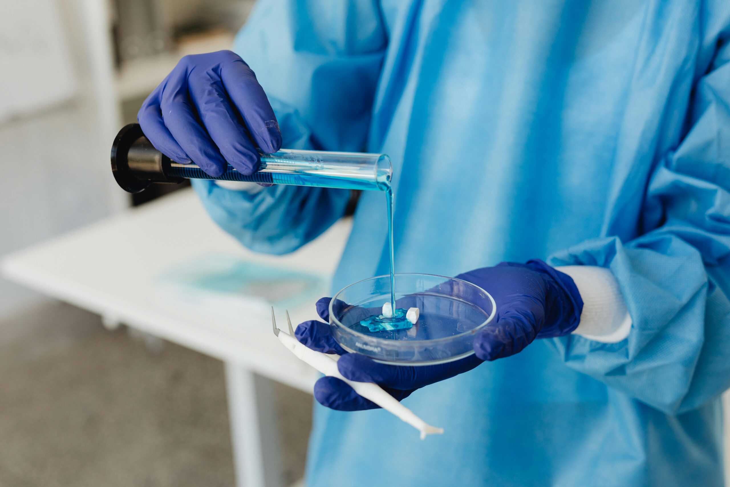 Online Classes Scientist in lab gown pouring blue liquid from test tube into petri dish wearing gloves.